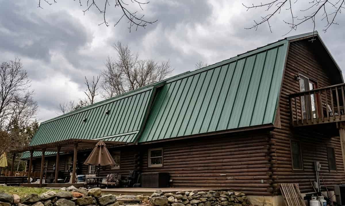 A wide, horizontal orientation view of a rustic log home featuring a large, green standing seam metal gambrel roof
