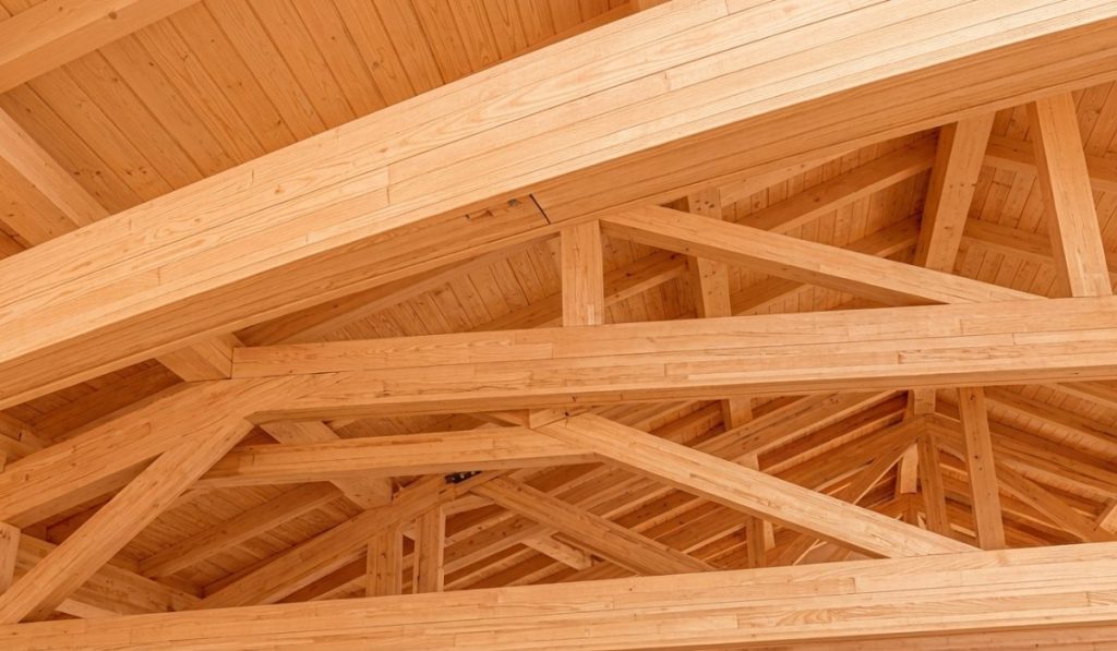 A close-up view of a complex, finished wooden ceiling structure showing interlocking light-colored timber beams and roof trusses