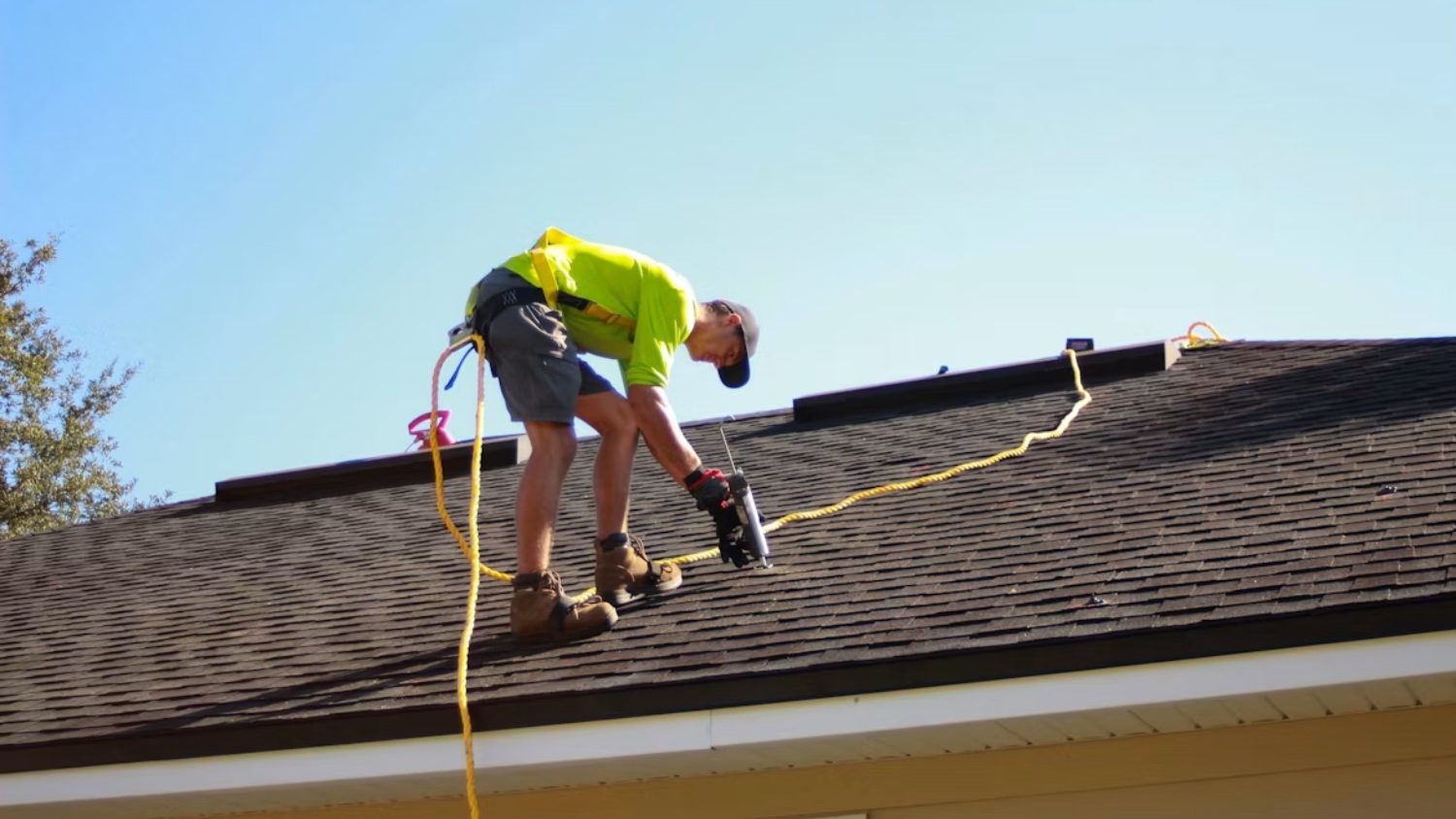 A roofer in a bright yellow safety shirt and harness uses a tool on a dark shingled roof