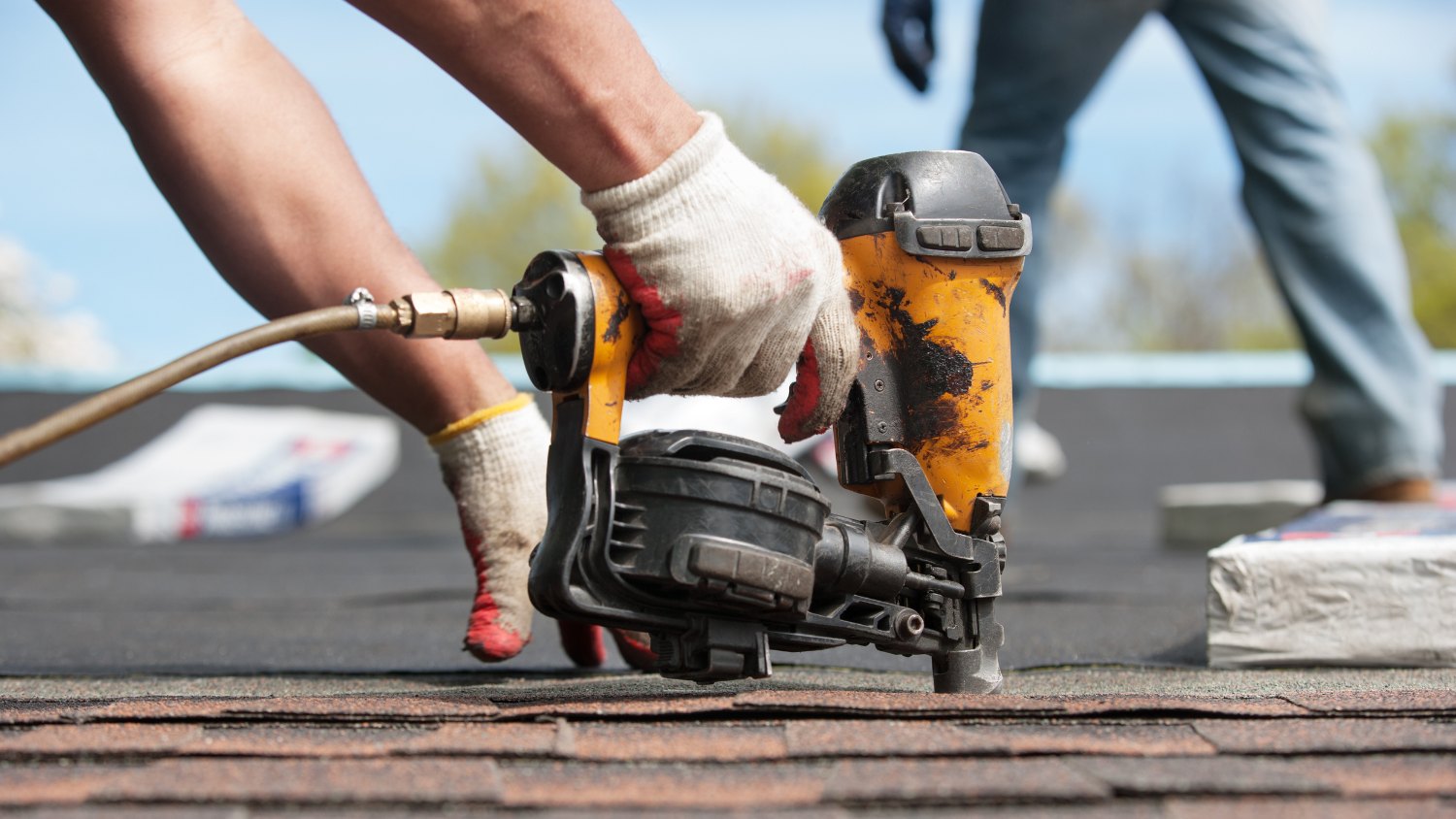 A roofer using a yellow pneumatic coil nail gun to install asphalt shingles