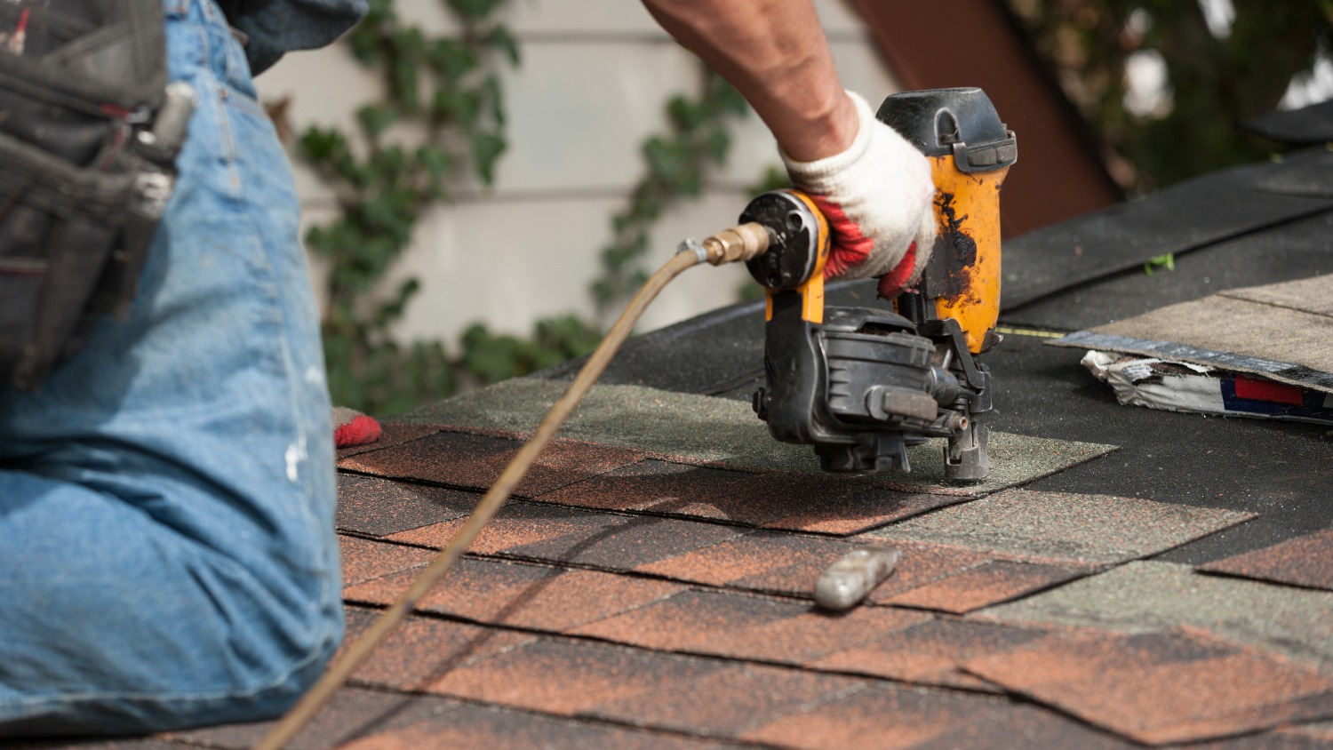 a worker using a pneumatic nail gun to install asphalt roof shingles