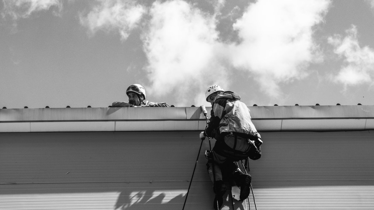 Two roofing contractors in safety gear inspecting rooftop drainage against cloudy sky